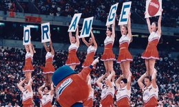 Female cheerleaders standing on male cheerleaders' shoulders and holding 7 signs that spell the word Orange with an orange fruit at the end