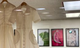 Two white dresses hanging behind glass in front of colorful framed pictures in Biblio Gallery