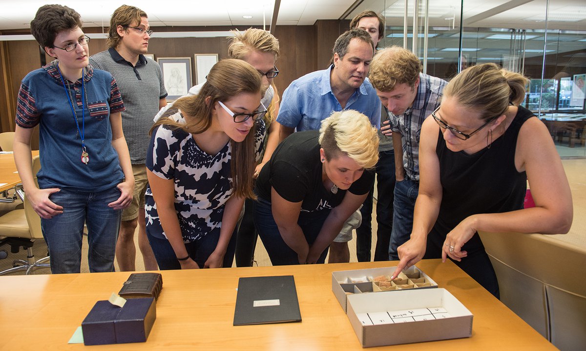 Students closely observing Nicolette Dobrowolski pointing toward a box of cuneiform tablets on a wooden table