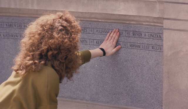 Archival image of a student in an olive green sweater kneeling and placing her hand on the Pan Am Flight 103 gray stone memorial wall engraved with names of victims